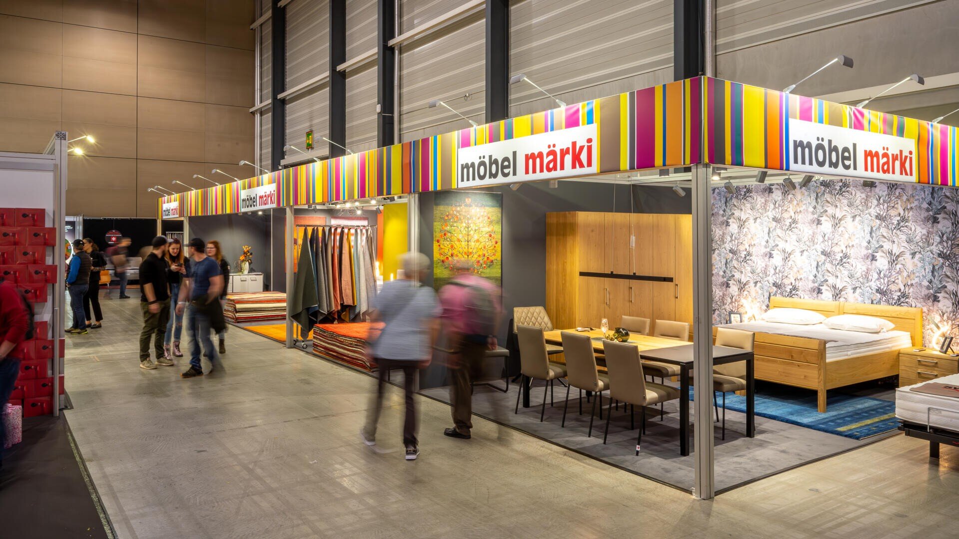 An einem Messestand mit der Aufschrift möbel märki werden Betten, Tische, Stühle und Schränke ausgestellt, die mit bunten Streifen beschriftet sind. Mehrere Leute gehen durch den Ausstellungsraum und schauen sich um.
