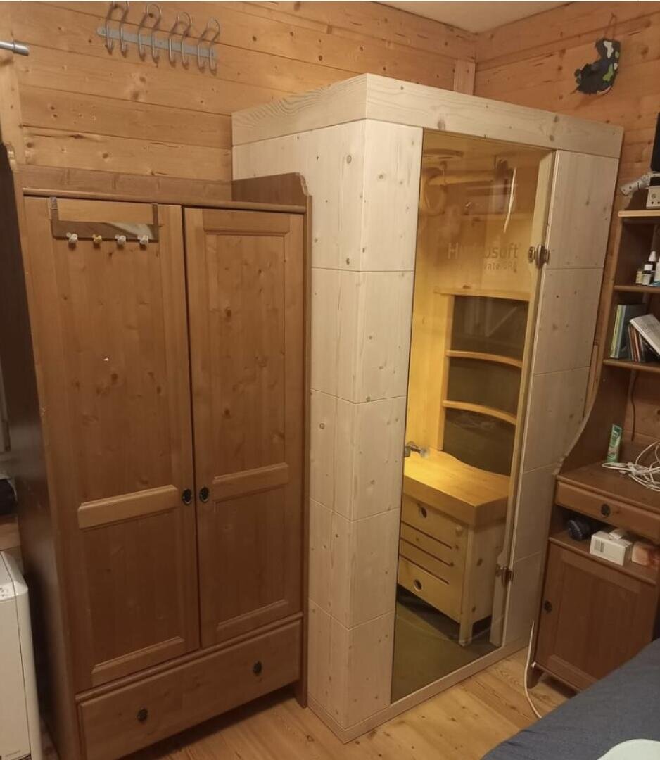Ein gemütliches Holzzimmer mit einem Kleiderschrank auf der linken Seite und einer modernen persönlichen Saunakabine mit Glastür und Innenbeleuchtung auf der rechten Seite, neben einem kleinen Schreibtisch mit Regalen.