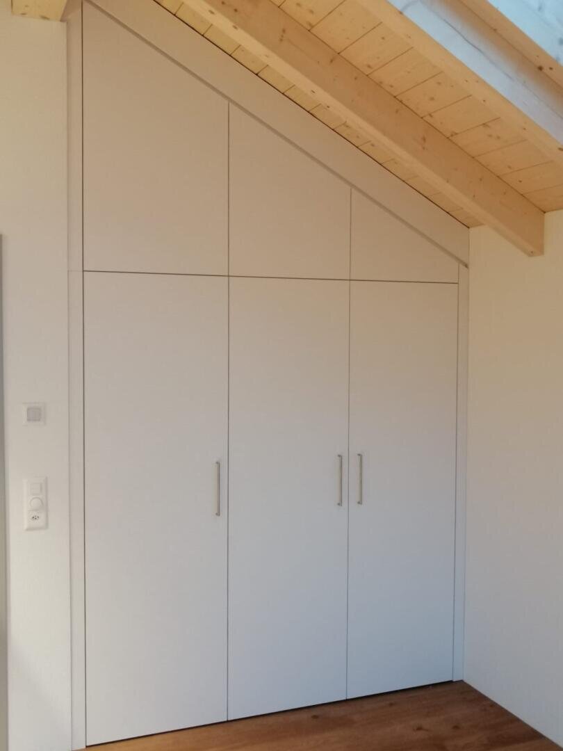 Weißer Einbauschrank mit drei Türen, jede mit einem vertikalen Griff, eingebaut unter einer schrägen Holzdecke in einem modernen, minimalistischen Raum mit Holzfußboden und weißen Wänden.