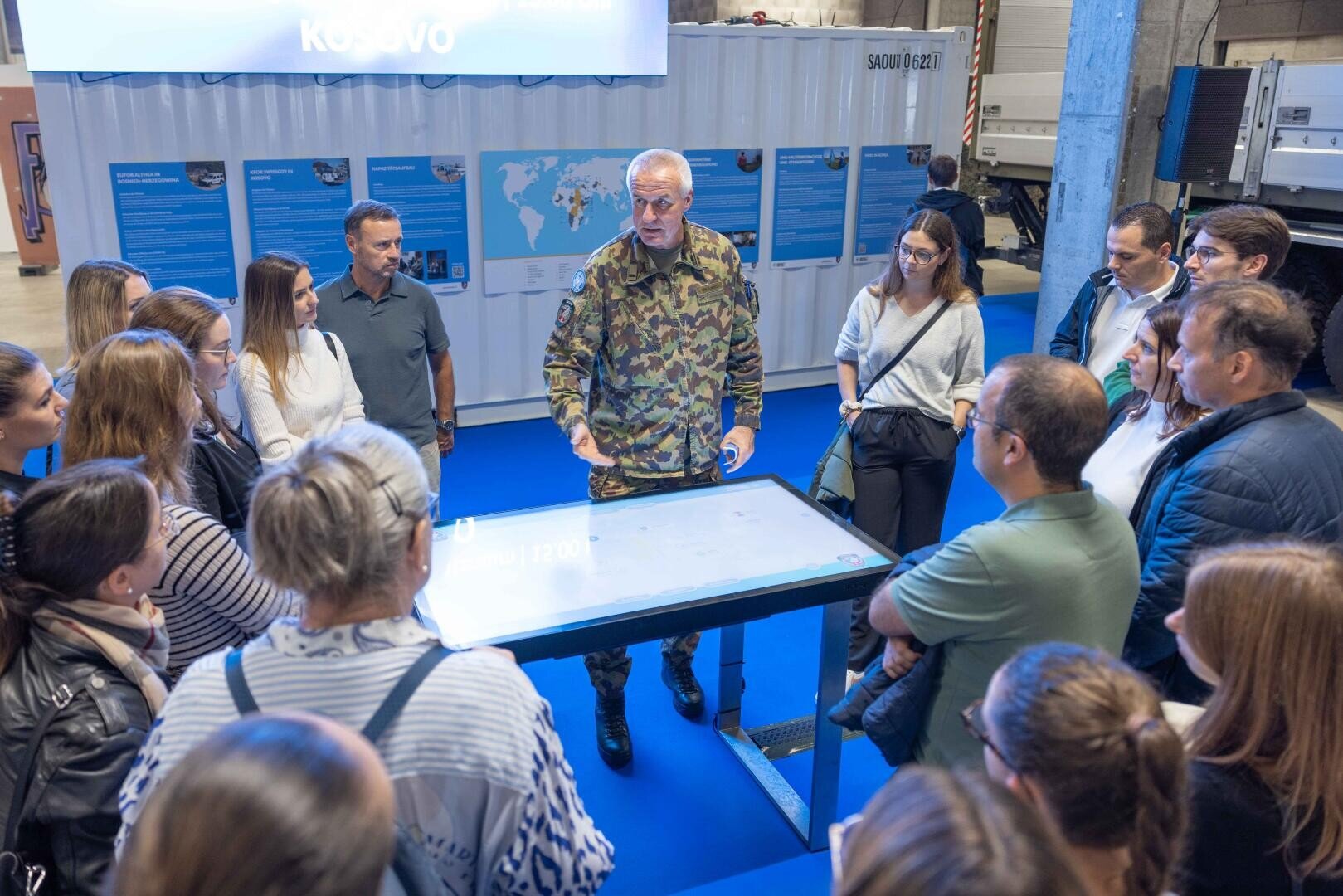 Ein Mann in tarnfarbener Militäruniform spricht zu einer Gruppe von Menschen, die sich in einem Innenraum um einen Tisch versammelt haben, während hinter ihm Informationsplakate an den Wänden hängen.