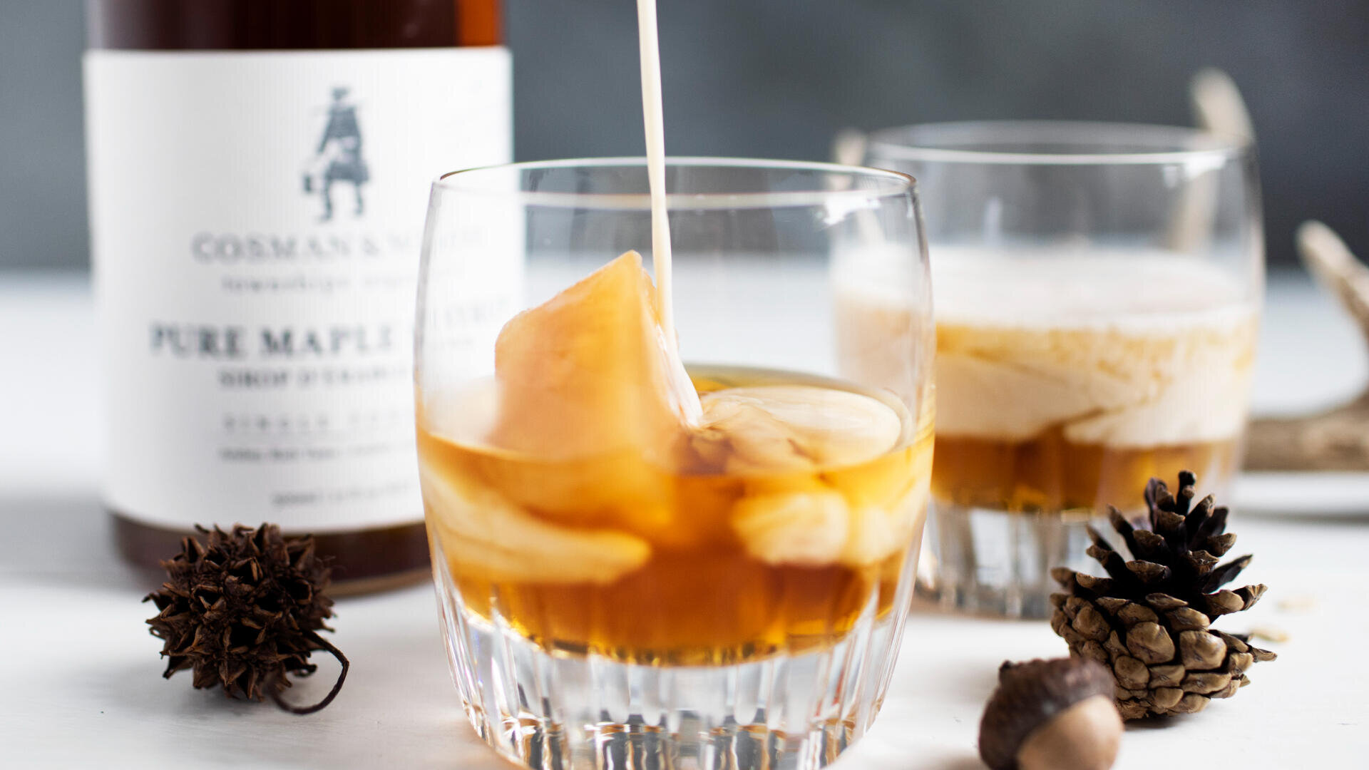 Eine Nahaufnahme von zwei Gläsern, die mit einem cremigen braunen Cocktail gefüllt sind, wobei eines mit Sahne aufgegossen wird. Im Hintergrund steht eine Flasche mit der Aufschrift "Pure Maple Syrup", und auf dem Tisch sind Tannenzapfen und Eicheln verstreut.