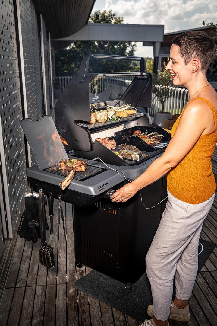 Eine Frau in einem ärmellosen orangefarbenen Oberteil grillt an einem sonnigen Tag verschiedene Fleisch- und Gemüsesorten auf einem Grill auf einer Holzterrasse und lächelt, während sie sich um das Essen kümmert.