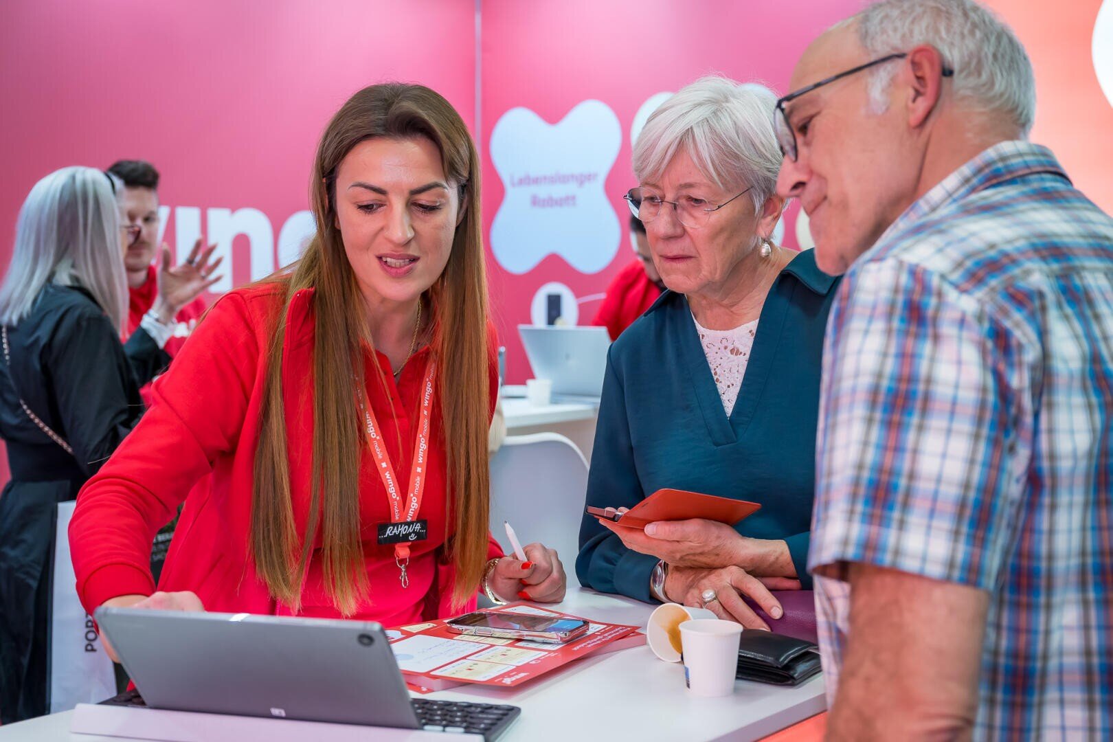 Eine Frau in einem roten Hemd zeigt einer älteren Frau und einem älteren Mann an einem Stand mit rosa Wänden Informationen auf einem Tablet. Die ältere Frau hält ein Telefon in der Hand und der Mann trägt eine Brille und ein kariertes Hemd. Im Hintergrund sind weitere Personen zu sehen.