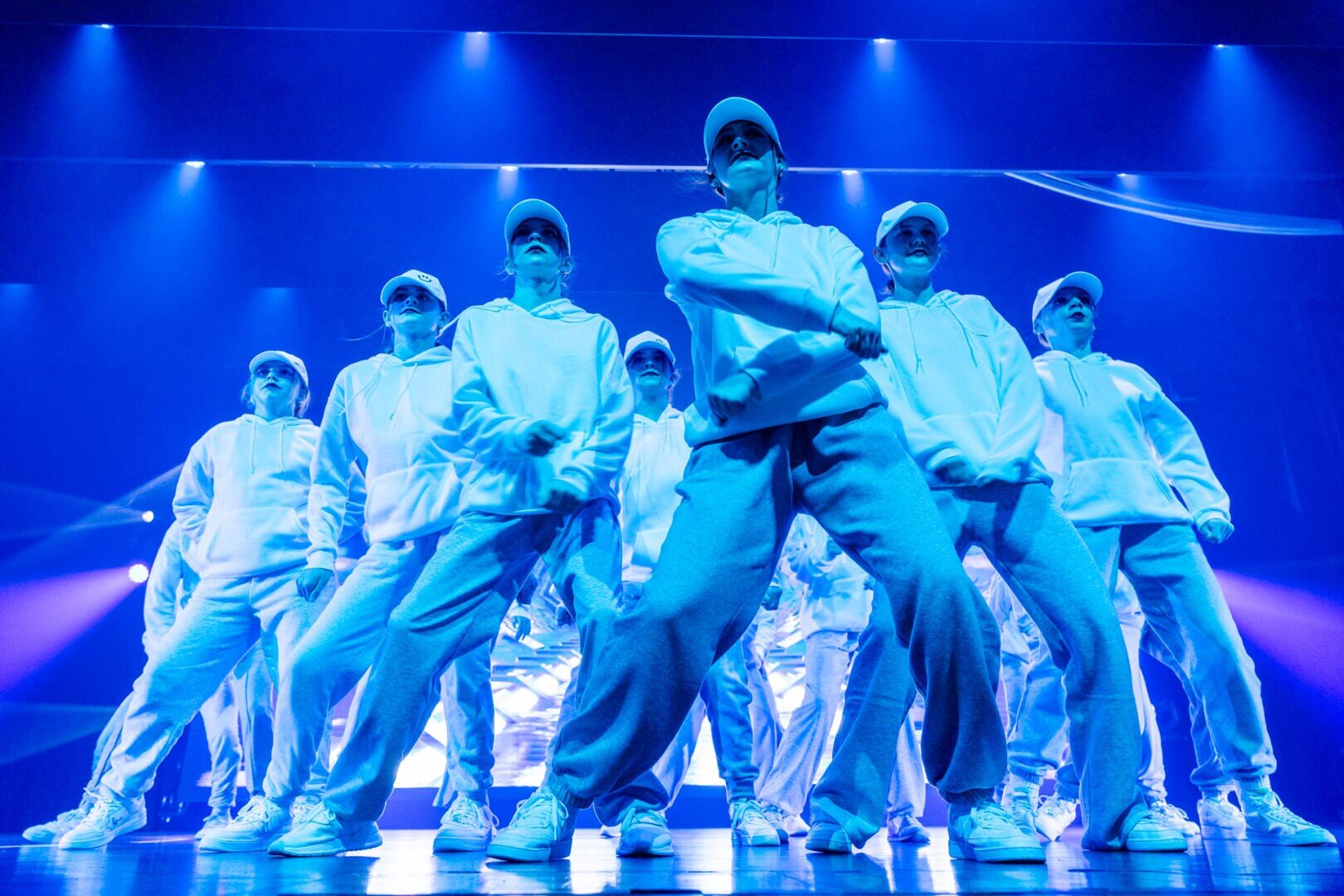 Eine Gruppe von Tänzern in passenden weißen Kapuzenpullis, Hosen und Mützen führt auf einer mit blauen und lila Lichtern beleuchteten Bühne eine Hip-Hop-Nummer auf.