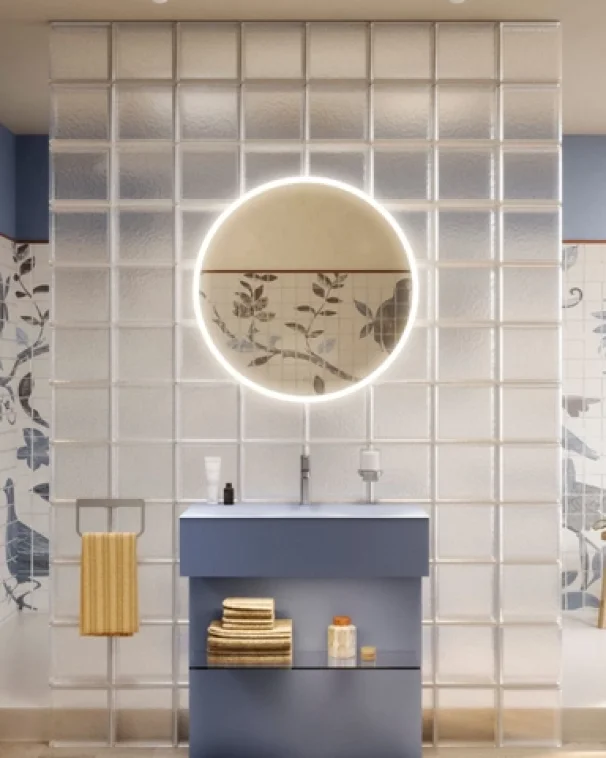Modernes Badezimmer mit einer Wand aus Glasbausteinen, einem runden, hinterleuchteten Spiegel über einem blauen Waschtisch und ordentlich gefalteten Handtüchern. An den Wänden sind Fliesen mit Blumenmuster zu sehen, und an einem nahegelegenen Handtuchhalter hängt ein gelbes Handtuch.