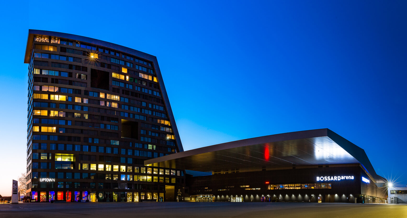 Ein modernes, geschwungenes Hochhaus mit vielen beleuchteten Fenstern steht neben der BOSSARD-Arena, beide in der Abenddämmerung vor einem tiefblauen Himmel beleuchtet. Die Gebäude haben ein schlichtes, zeitgenössisches Design.