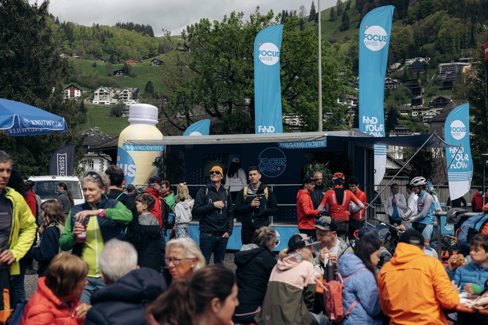 Eine große Menschenmenge, viele mit Jacken und Outdoor-Ausrüstung, versammelt sich bei einer Veranstaltung im Freien mit blauen FOCUS WATER-Bannern und einem Promo-Stand vor einer Kulisse aus grünen Hügeln und Häusern.